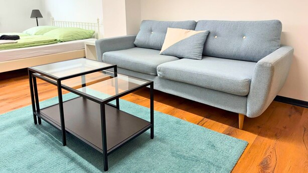 A room with a grey sofa, a glass-top coffee table, a bed with green bedding, and a small table with a lamp. - House with free public parking (Köln)