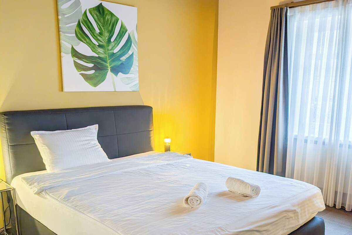 A hotel room with a large bed, a headboard, a wall-mounted artwork, a bedside lamp, and a window with curtains.