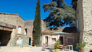 A courtyard with stone buildings, a tall tree, outdoor seating, and a green umbrella. - LE MAS GORCE, Drôme valley, spacious TERRACE, courtyard, parking, garage. (Chabrillan)