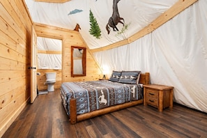 Una habitación tipo tienda con suelo de madera, una cama con ropa de cama estampada, una mesita de noche de madera, un espejo y un inodoro.