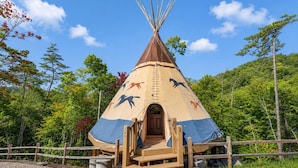 Un tipi doté d’un porche en bois et d’une échelle, entouré d’une clôture en bois, situé dans une zone boisée.