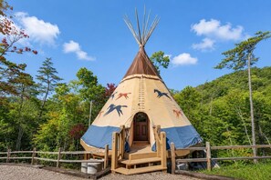 Un tipi con un porche de madera y una escalera, rodeado por una cerca de madera, situado en una zona boscosa.