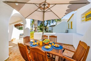 An outdoor dining area with a table set for two, surrounded by wooden chairs, under a large umbrella.
