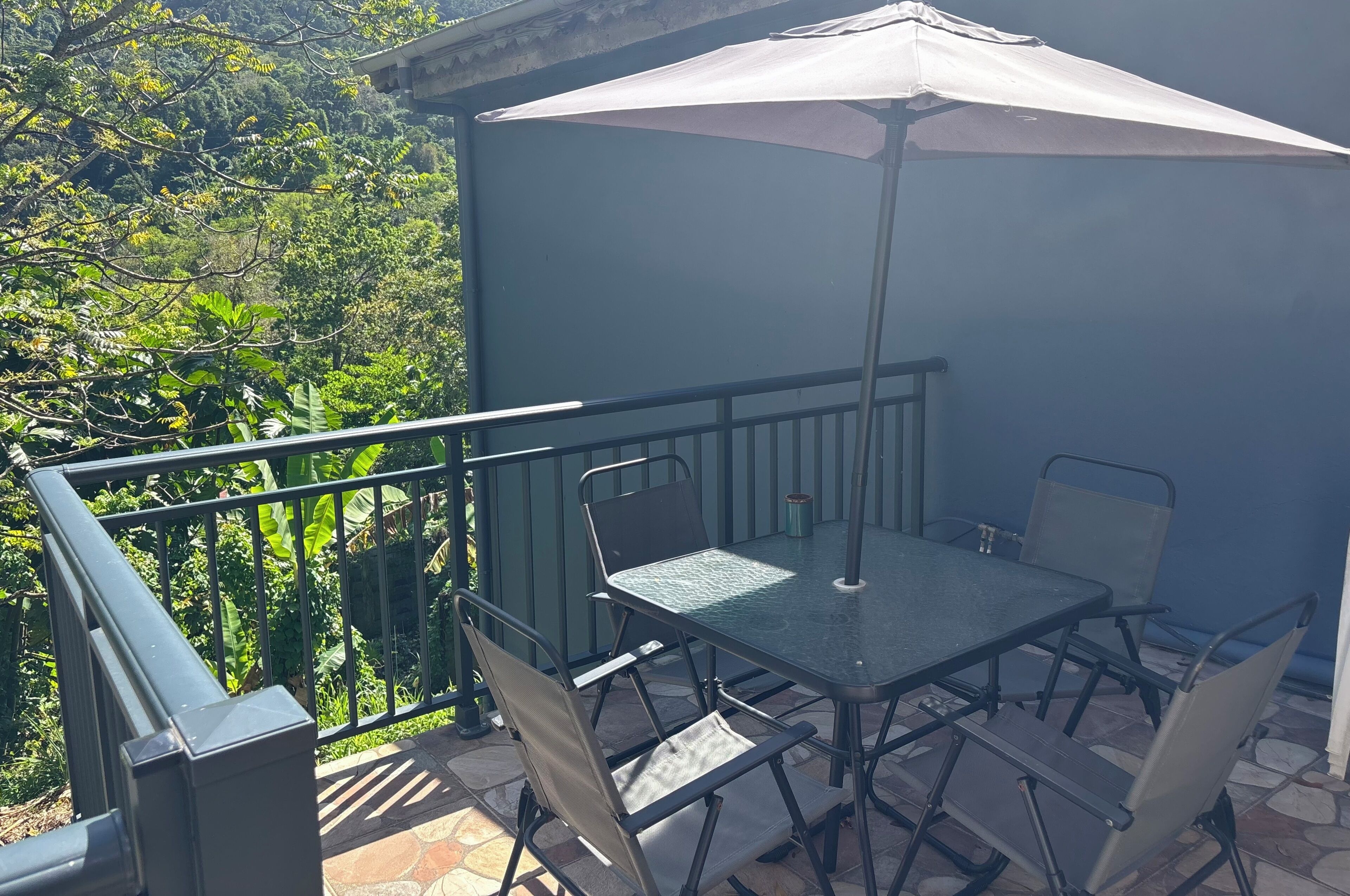 An outdoor patio with a table and chairs, a large umbrella, and a view of greenery.