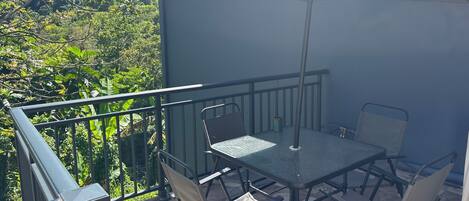 An outdoor patio with a table and chairs, a large umbrella, and a view of greenery.