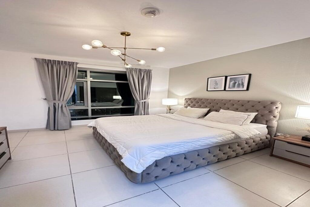 Ein Schlafzimmer mit einem großen Bett, einem Fenster mit Vorhängen, einem Nachttisch und einer Wandleuchte.