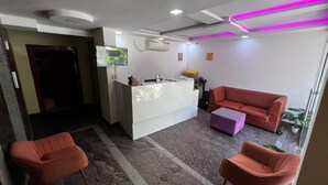 Uma área de recepção com uma mesa branca, um sofá vermelho, duas cadeiras vermelhas, um banquinho roxo e iluminação roxa no teto.