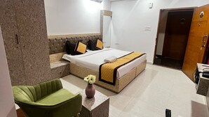 Quarto de hotel com uma cama, uma cadeira verde, uma pequena mesa com um vaso, um ventilador de teto e uma porta de madeira.