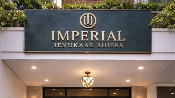 Entrada do Imperial Jenukaal Suites com design moderno, apresentando uma placa grande, degraus que levam à entrada e plantas em vasos de ambos os lados.