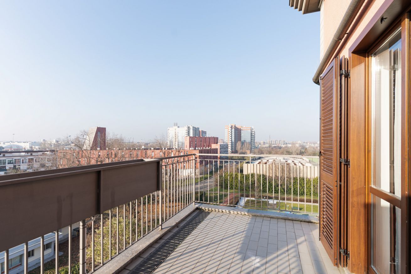 Ein Balkon mit Metallgeländer, einer Holztür und Blick auf Gebäude und Grünflächen.