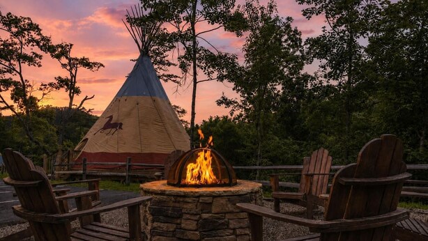 Ein Tipi mit Feuerstelle davor, umgeben von Holzstühlen.