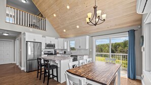 A modern kitchen with white cabinets, stainless steel appliances, a wooden dining table, and a balcony with a view of greenery.