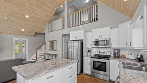 A modern kitchen with white cabinets, stainless steel appliances, a marble countertop, and a wooden ceiling.