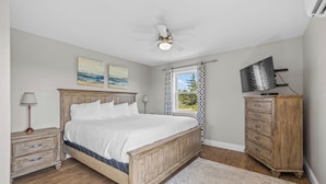 A bedroom with a wooden bed, a nightstand, a dresser, a ceiling fan, a TV, and a window with curtains.