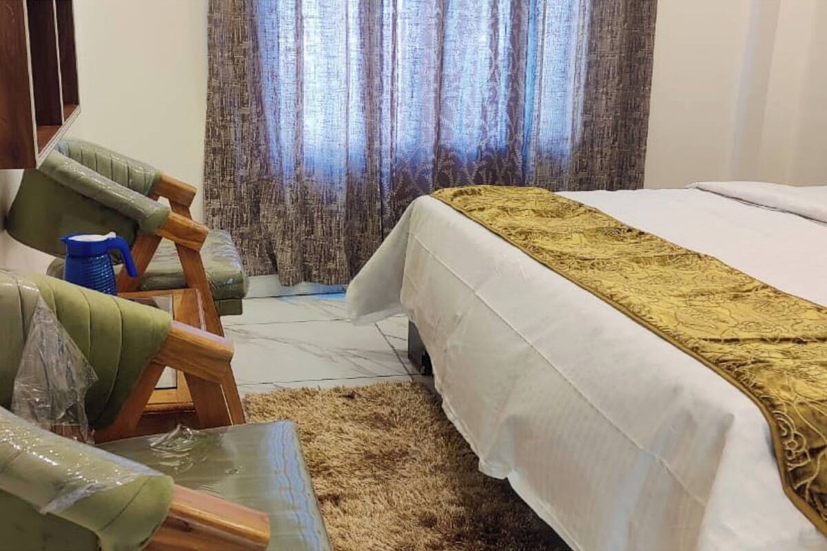 Une pièce comprenant un lit, un fauteuil vert, une chaise bleue, une petite table et un rideau.