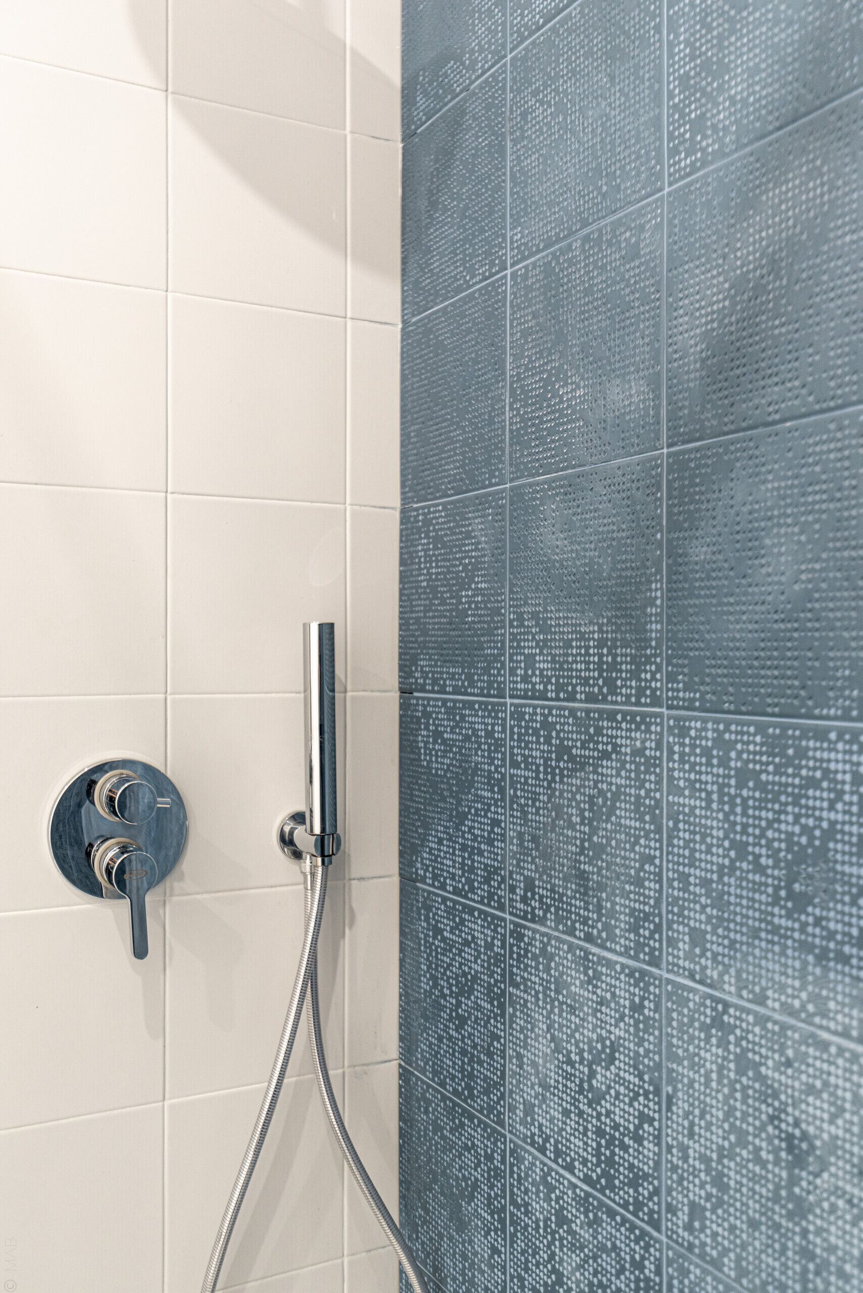 Un espace douche doté d’un robinet et d’un flexible chromés, de murs carrelés dans deux nuances de couleurs et d’une molette de réglage.