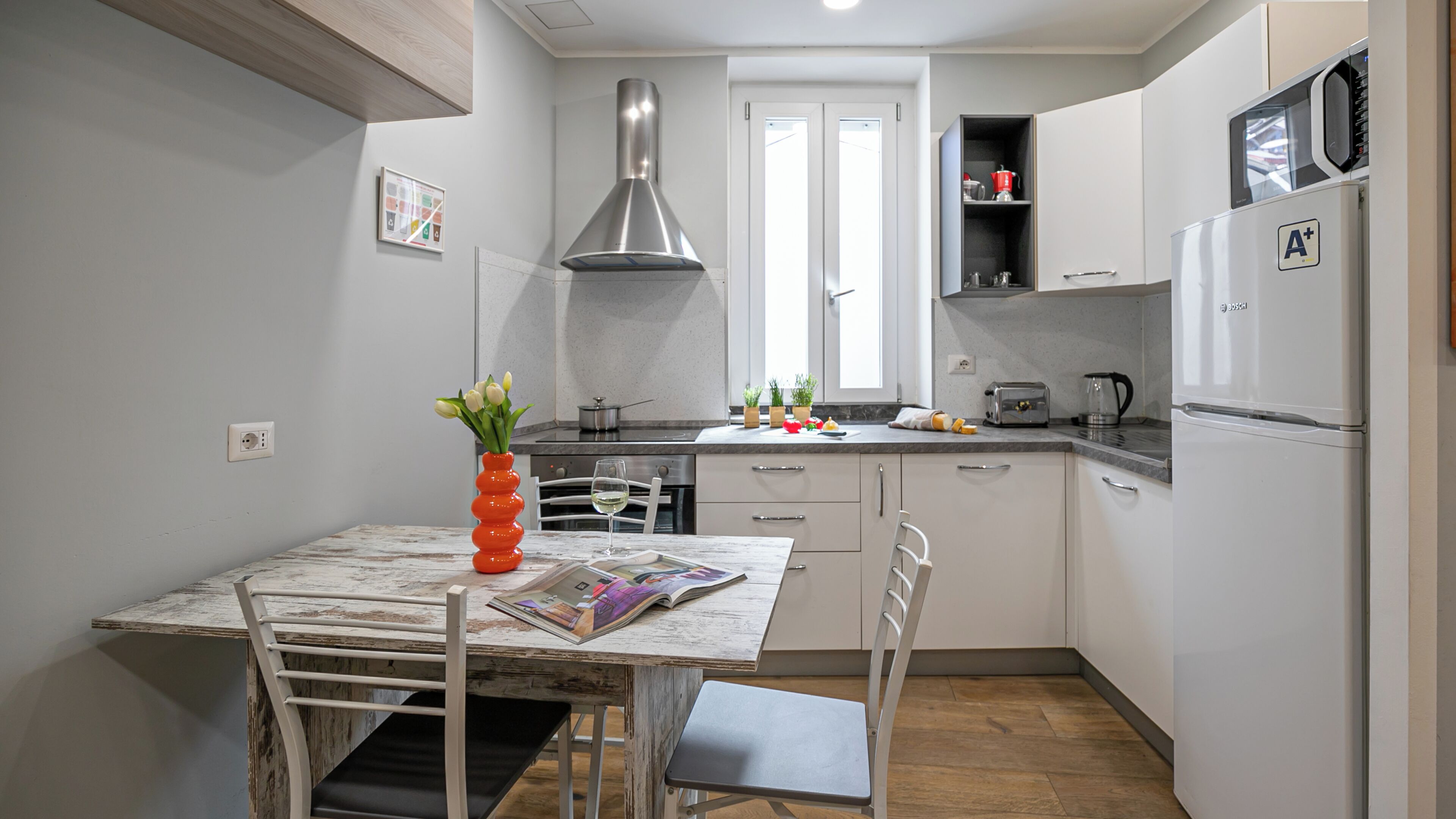 A compact kitchen with a dining table, chairs, a red vase with flowers, a window, and a Bosch refrigerator.