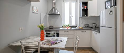 A compact kitchen with a dining table, chairs, a red vase with flowers, a window, and a Bosch refrigerator.