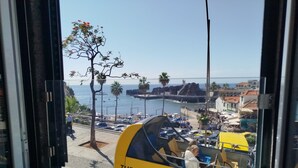 Une fenêtre de bus donnant sur une zone côtière, avec vue sur une plage, un promontoire rocheux et un bâtiment doté d’un toit rouge.