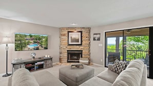 A living room with a stone fireplace, a television mounted on the wall, and a balcony with a view of trees. - Lake Ozark Oasis Central Location & Pool Access (Lake Ozark)