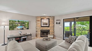 A living room with a stone fireplace, a television mounted on the wall, and a balcony with a view of trees. - Lake Ozark Oasis Central Location & Pool Access (Lake Ozark)