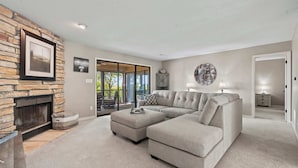 A living room with a stone fireplace, a sectional sofa, a round wall clock, and a view through a glass door to an outdoor patio. - Lake Ozark Oasis Central Location & Pool Access (Lake Ozark)