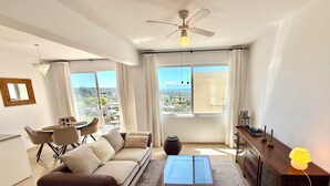 Un soggiorno con un divano, un tavolino, un mobile TV, una ventola a soffitto e vista sulla città attraverso ampie finestre.