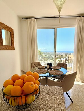 Un coin repas avec une table en verre, des sets de table en fibres tressées et un saladier rempli d’oranges.
