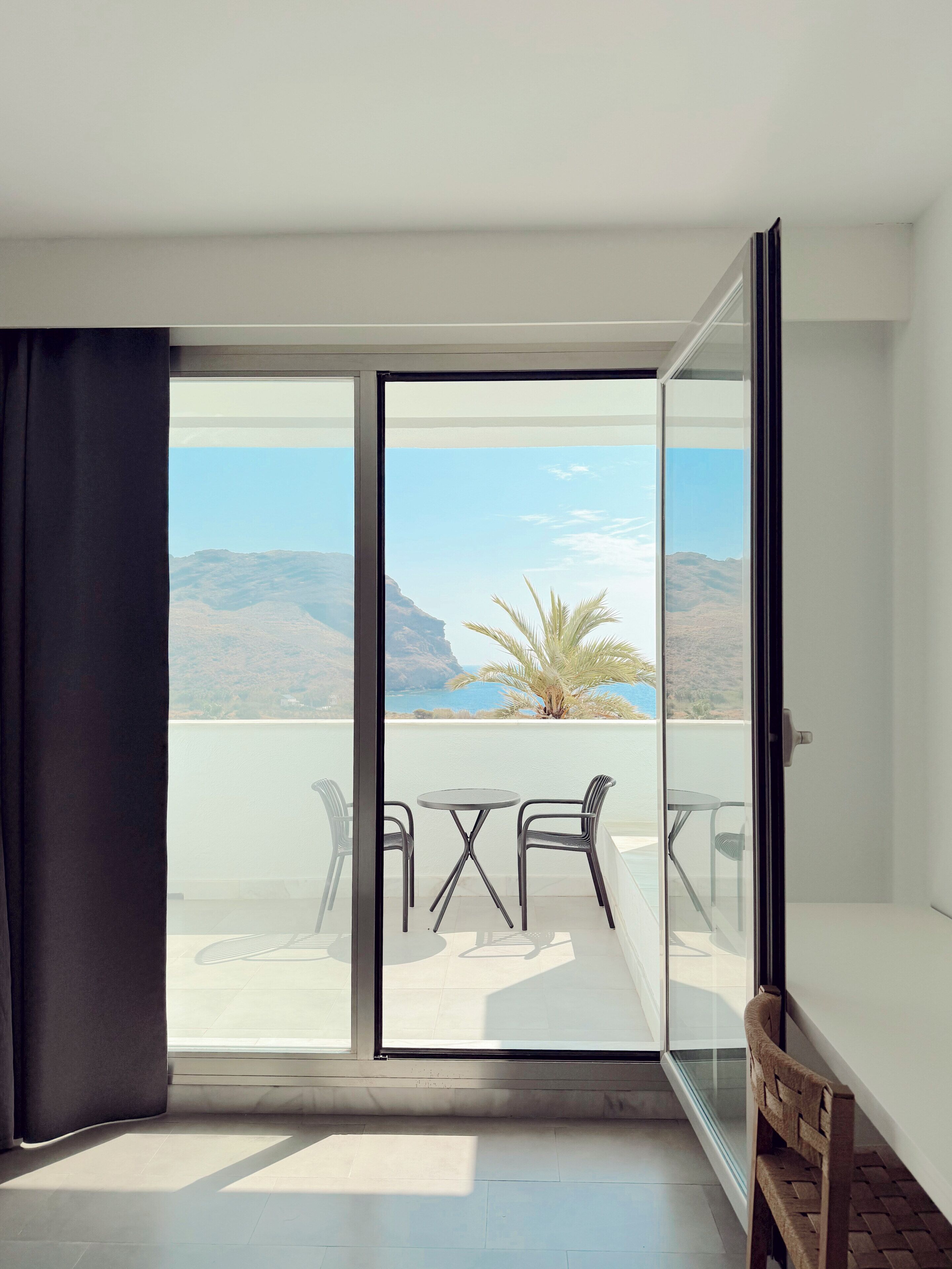 A balcony with a table and chairs, a view of the sea and mountains, and a sliding glass door.