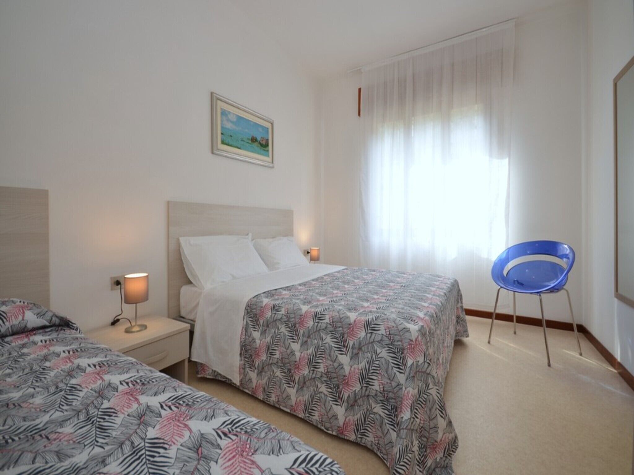 A hotel room with a bed, a chair, a nightstand, a lamp, and a window with sheer curtains.