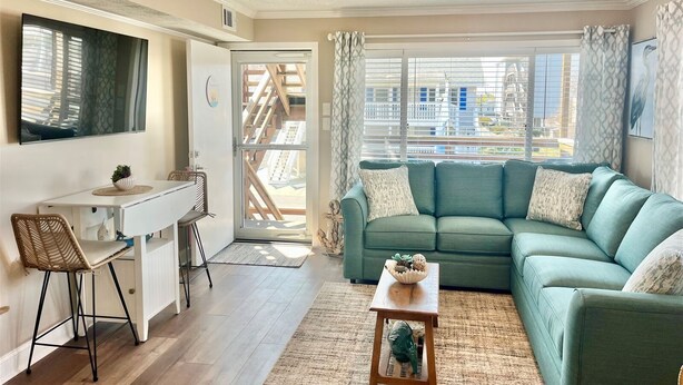 A modern living room with a green sofa, a dining table with chairs, a small side table, and a large window with a view of the outside. - New for 2026! Ocean Block and Fully Renovated! Comfortably sleeps 6, FREE Linens (Ocean City)