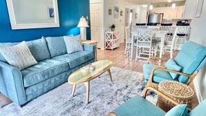 Ein Wohnzimmer mit einem blauen Sofa, einem Couchtisch und einem Essbereich mit weißen Stühlen, im Hintergrund eine Küche.