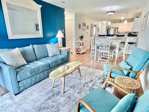 A living room with a blue sofa, a coffee table, and a dining area with white chairs and a kitchen in the background. - New 2026! Bayfront, FREE Linens & Towels, Outdoor Pool, Walk to Fager's Island (Ocean City)