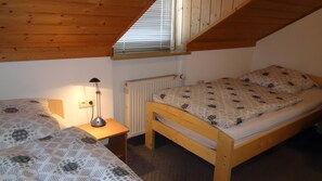 Une petite pièce agréable, dotée de deux lits en bois, d’une table de chevet avec une lampe, d’un radiateur et d’un plafond en pente.