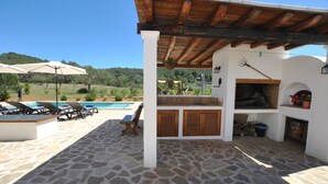 A covered outdoor area with a stone floor, a pool, lounge chairs, and a barbecue grill. - Charming villa in the middle of nature (Sant Miquel de Balansat)