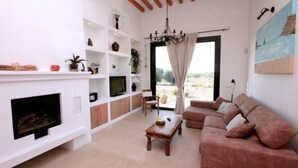A living room with a fireplace, sofa, TV, and a view through a large window. - Charming villa in the middle of nature (Sant Miquel de Balansat)