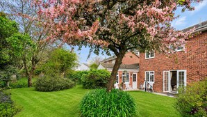 Un jardín bien cuidado con césped, un árbol con flores rosadas y una casa de ladrillo con un patio visible.