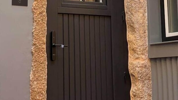 Eine dunkelbraune Tür mit Fenster, begrenzt von strukturierten Steinwänden, und einer kleinen Lampe darüber.