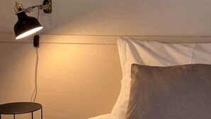 Ein Bett mit weißer Bettwäsche und einem grauen Kopfkissen, eine schwarze Schreibtischlampe auf einem Nachttisch und ein gerahmtes Bild eines Gebäudes an der Wand.