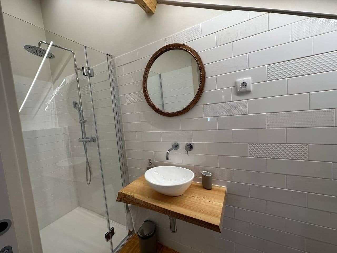 A modern bathroom with a glass shower enclosure, a wooden vanity with a white sink, and a round mirror.
