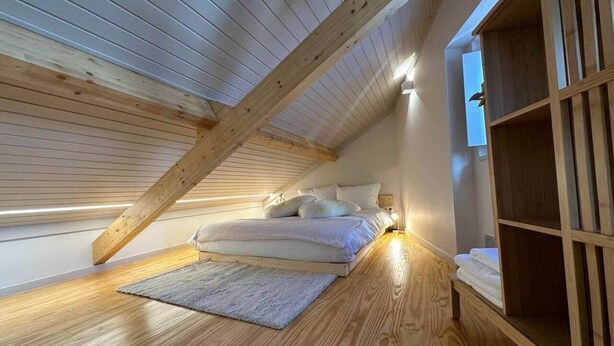 Un dormitorio con techo inclinado, suelo de madera, cama con almohadas, mesita de noche y estante de madera empotrado.