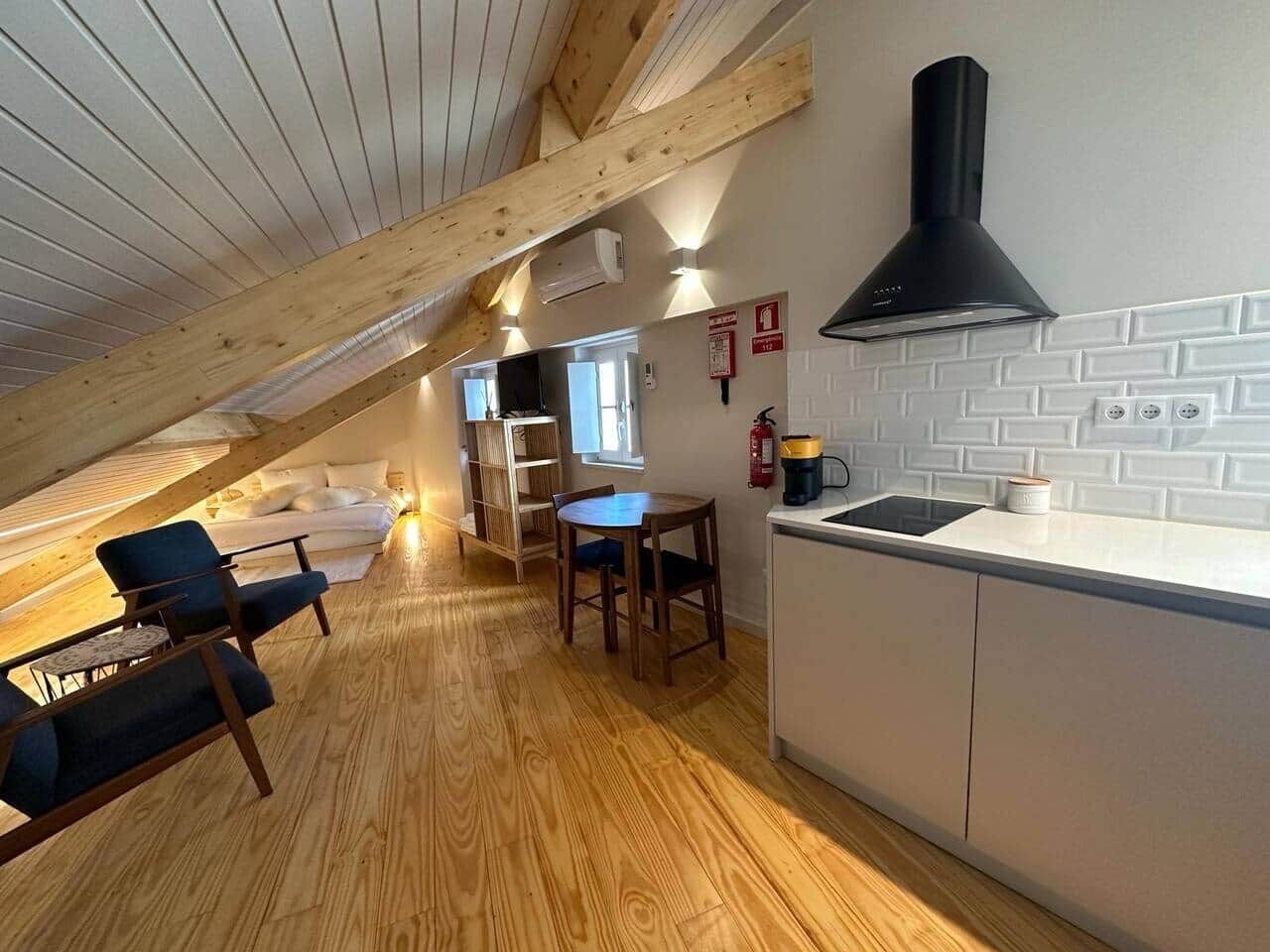 An attic bedroom with a wooden ceiling, a kitchenette, a dining area with a table and chairs, and a sofa.