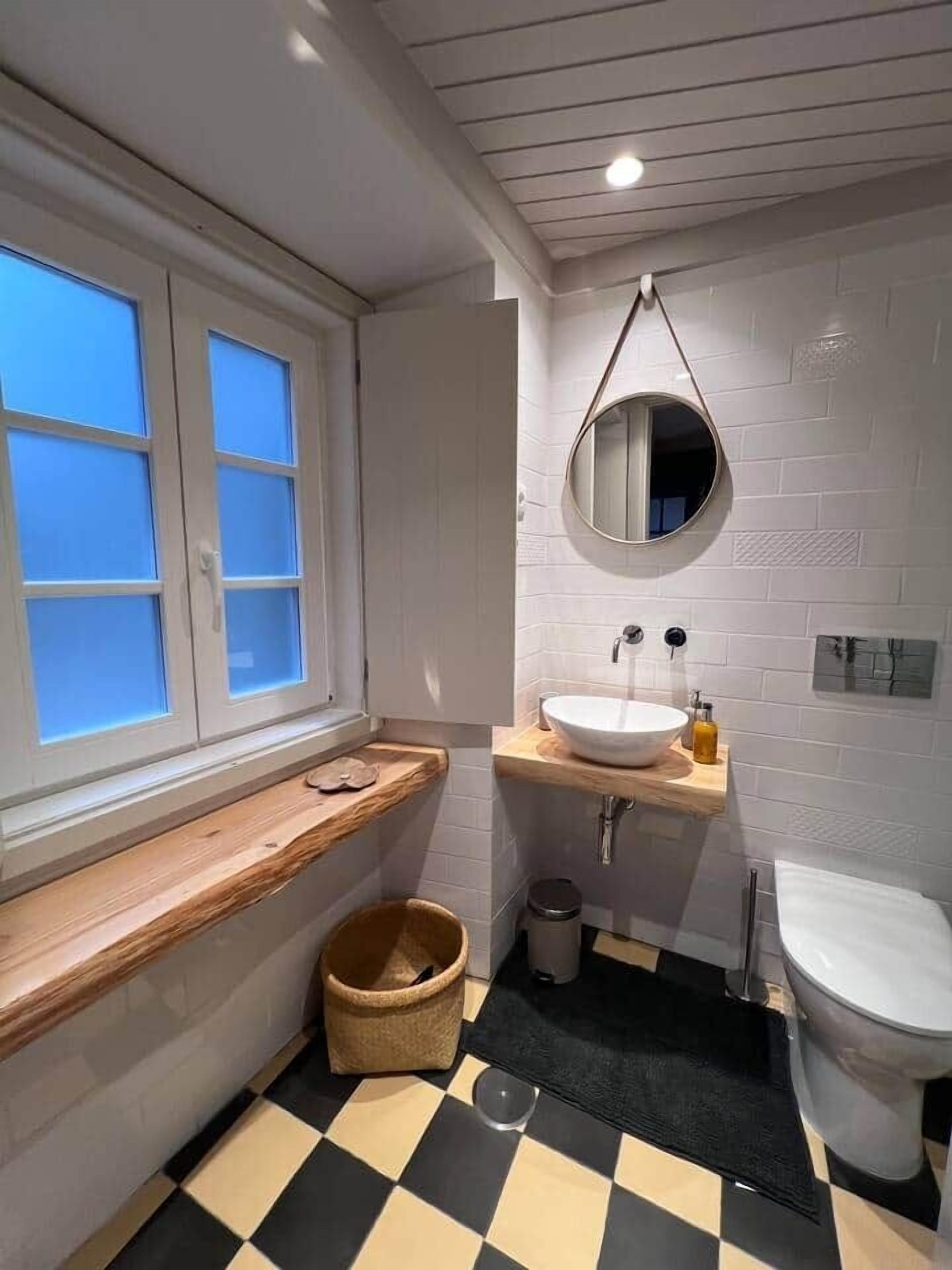 A bathroom with a window, a sink, a mirror, a toilet, and a checkered floor.
