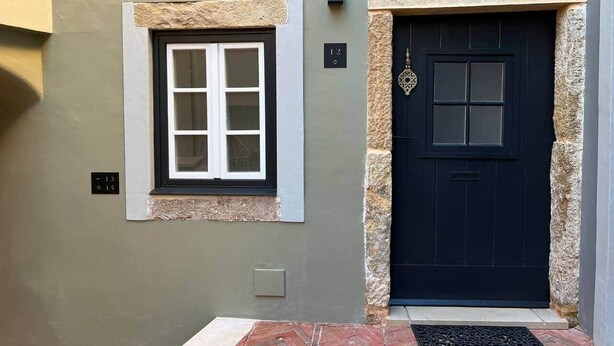A building with a black door, a window with white framing, and a stone accent around the door. - CorpusChristi 35-12 (Coimbra)