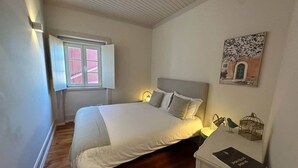 Quarto com cama, mesa de cabeceira, quadro na parede e janela com persianas.
