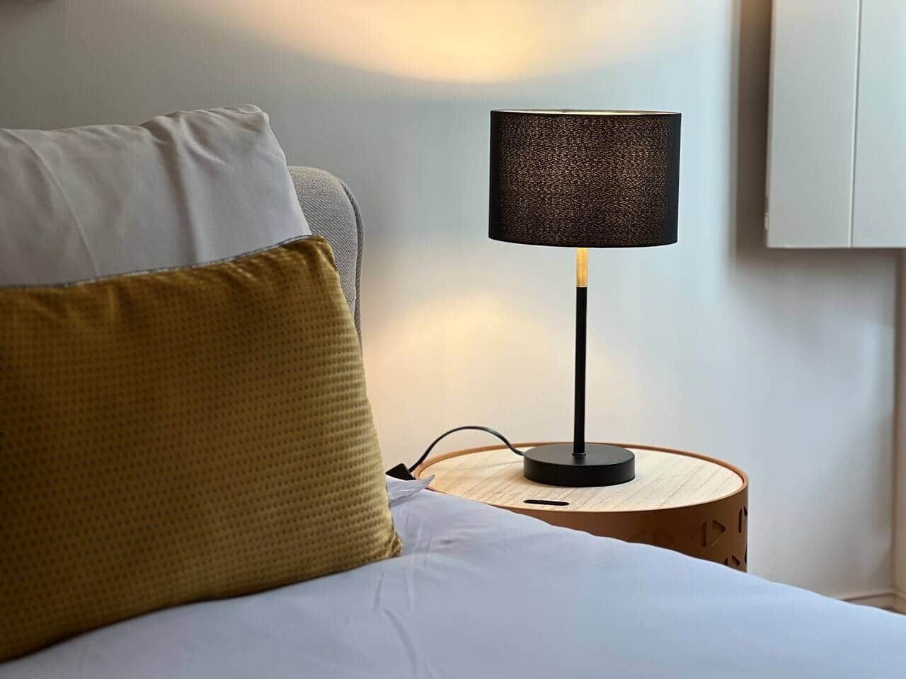 Un lit avec des oreillers blancs et moutarde, une table de chevet ronde en bois avec une lampe noire, et un mur blanc.