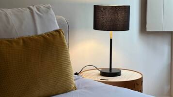 Un lit avec des oreillers blancs et moutarde, une table de chevet ronde en bois avec une lampe noire, et un mur blanc.
