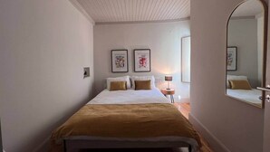 Ein Schlafzimmer mit einem Bett, einem Nachttisch, einer Lampe, zwei gerahmten Bildern an der Wand und einem Spiegel.