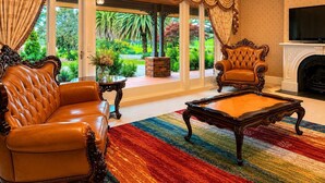 A spacious living room with a leather sofa, a matching armchair, a coffee table, and a fireplace. Large windows offer a view of a lush garden. - Oak Hill Grand Country Escape with Sunset Views (Chirnside Park)