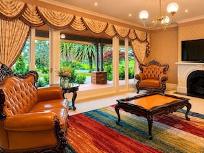 A spacious living room with a leather sofa, a matching armchair, a coffee table, and a fireplace. Large windows offer a view of a lush garden.
